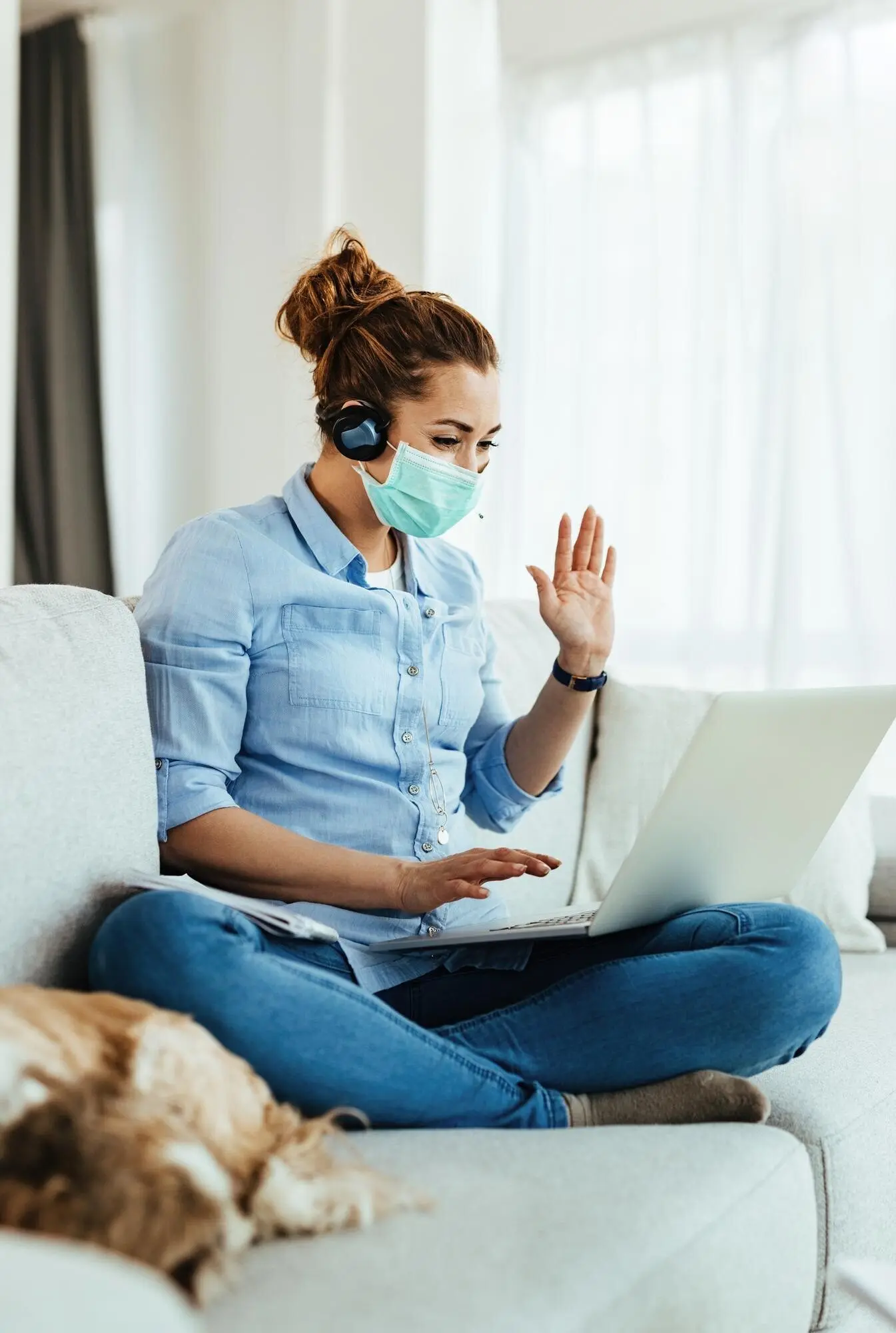 A happy woman wearing a protective face mask is using a laptop and waving to someone while making a video call from home.