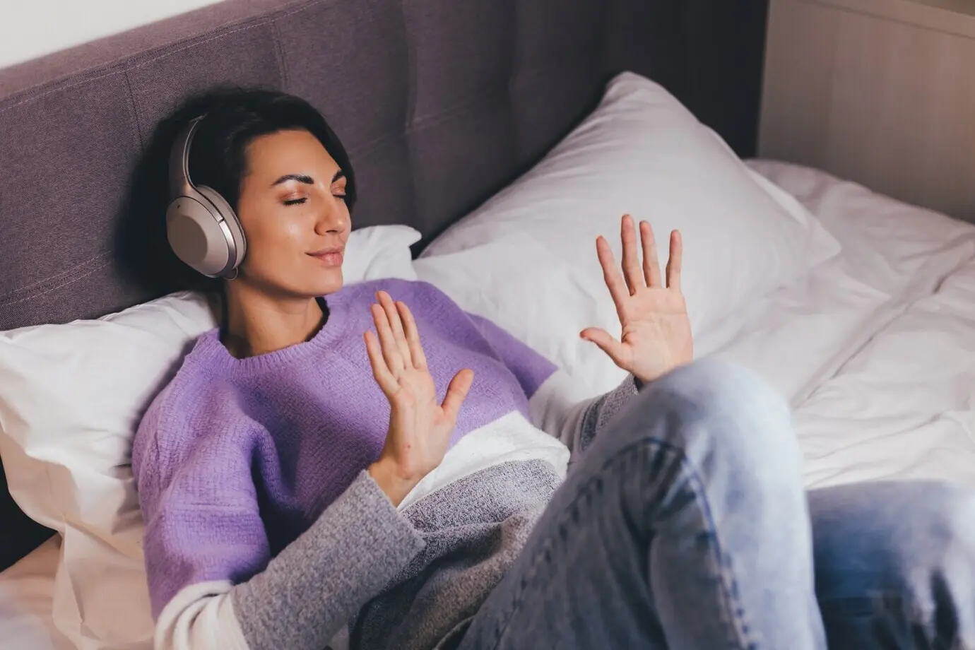 A happy woman at home on a comfy bed, wearing a warm pullover and listening to music.